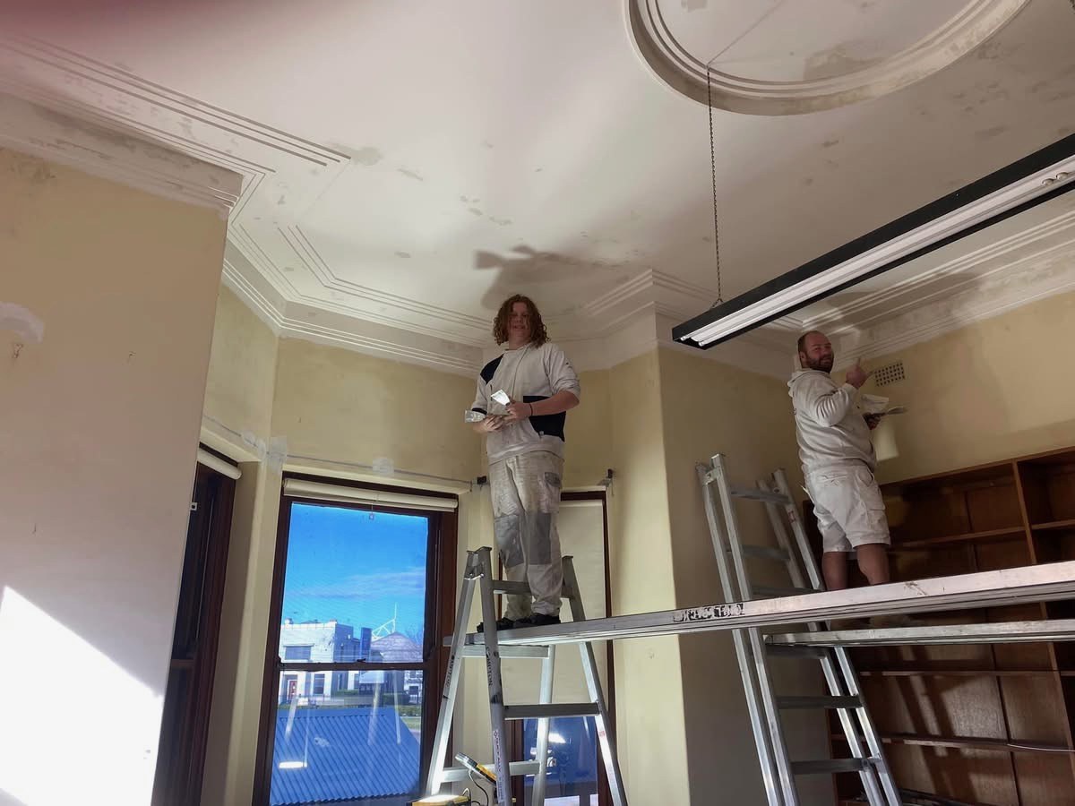 Before: Heritage room before restoration, with painters on ladders preparing the ceiling and cornices